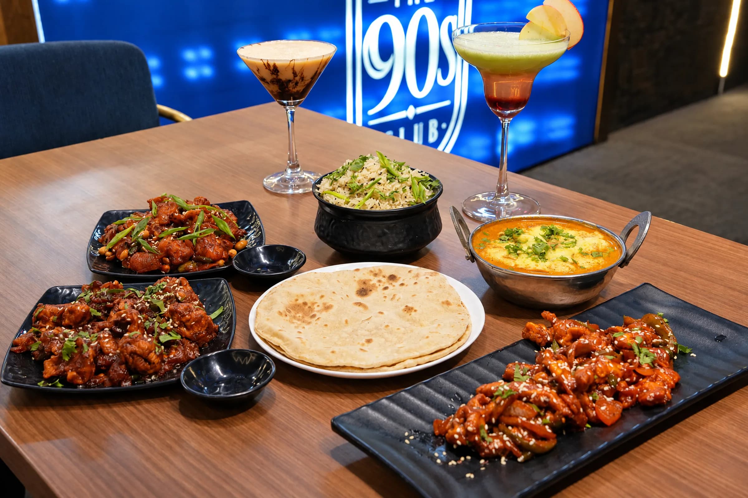 Chilli chicken, a bowl of biryani, a curry, flatbread and two cocktails on a table at The 90s Club in Electronic City, with the lit bar logo behind.