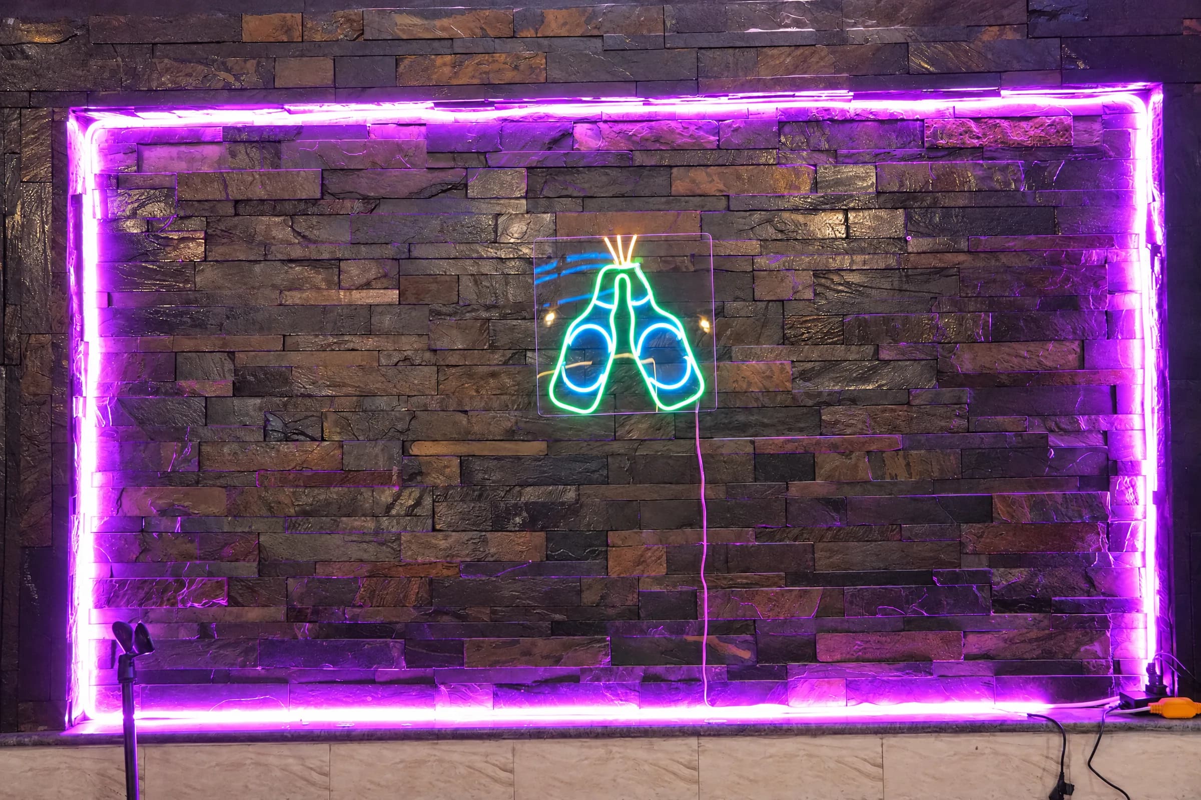 A neon sign of two clinking beer bottles on a stone wall framed in purple light at The 90s Club.