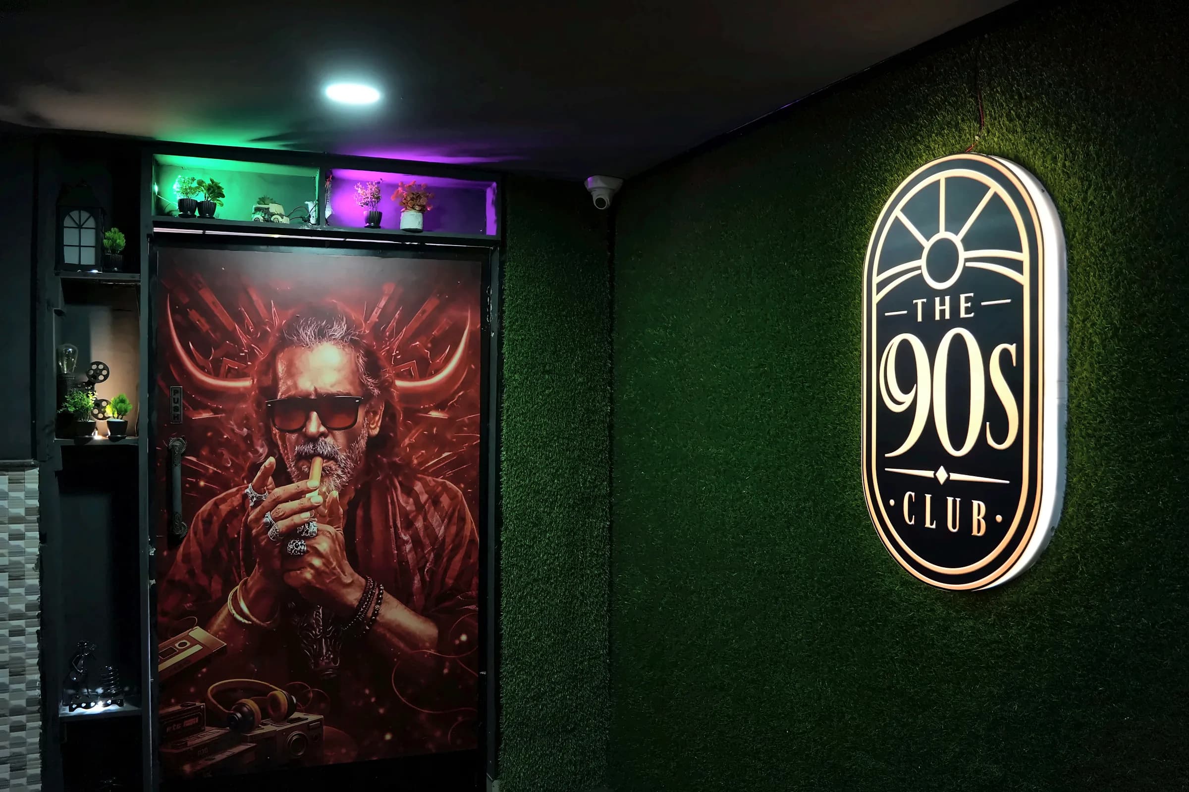 The backlit 90s Club logo on a green wall beside a large portrait artwork inside The 90s Club, Electronic City.